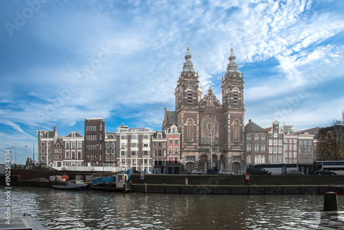 Historic Amsterdam cityscape with the Basilica of Saint Nicholas prominently featured. The scene is under a bright blue sky with scattered clouds