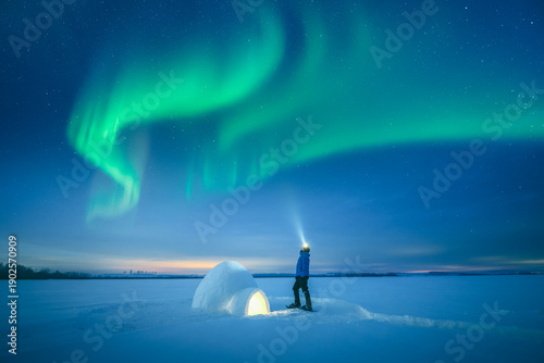 Polar survival concept with man using flashlight near illuminated snow igloo in open winter field, aurora borealis, arctic night scene, polar lights, northern lights