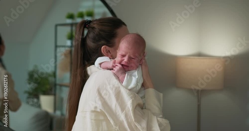 Mother cradles her newborn baby on her shoulder in a softly lit room with a lamp and mirror in the background. Mother Holding Newborn Baby