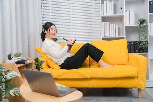 Fotografía Home Office Bliss: A young woman, radiating tranquility, comfortably reclines on a bright yellow sofa, immersed in her work, a laptop at her side, capturing the harmony of work-life integration