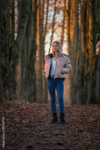 Young woman in a beige puffer jacket and blue jeans stands confidently on a leaf-covered path surrounded by tall trees in a serene forest setting