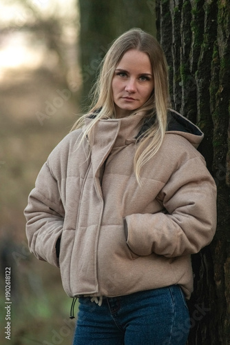 Young woman in a beige puffer jacket and blue jeans stands confidently on a leaf-covered path surrounded by tall trees in a serene forest setting