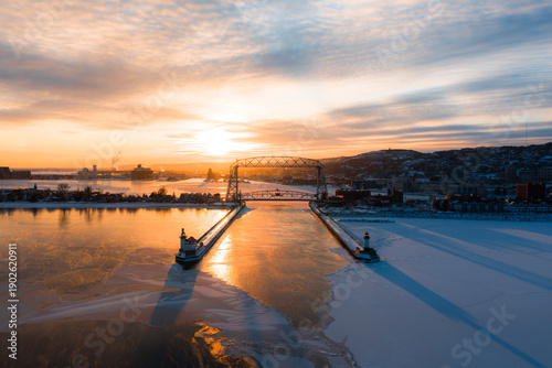 A wide aerial perspective of Duluth's Canal Park, the Aerial Lift Bridge, and the lighthouses under a brilliant winter sunrise.