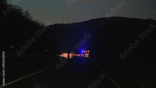 View from a car of a highway road block at dusk. Flashing blue emergency lights and reflective barriers signal a warning and detour. Dark, moody, and cautious atmosphere with lens flare