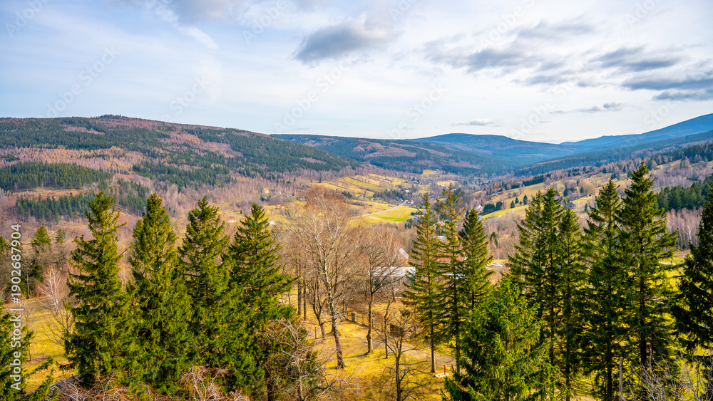 Fototapeta premium A wide view of Swieradow Zdroj in Poland shows rolling hills and trees under a blue sky. The landscape has patches of grass and areas of bare trees. Mountains rise in the distance.