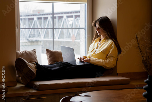 Young woman working on laptop by a cozy window seat