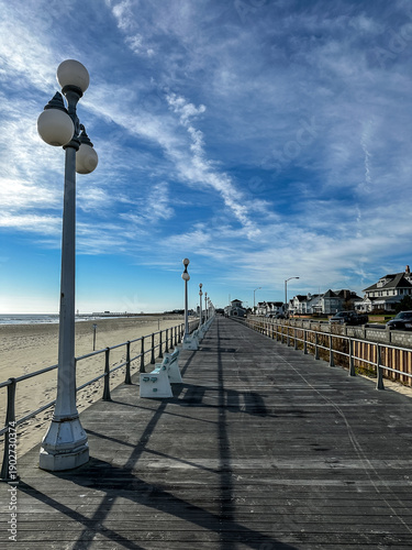 Avon by the Sea, New Jersey - Wooden boardwalk stretching alongside a sandy beach under a blue sky with scattered clouds. The boardwalk is flanked by vintage-style street lamps and a railing
