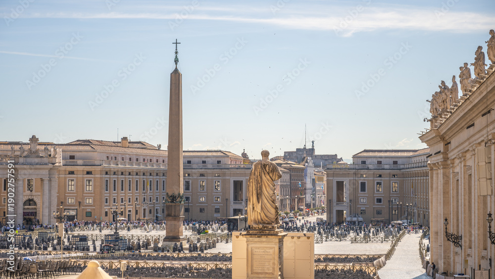 Naklejka premium Visitors walk through St Peter's Square in Vatican City. The sun shines over the large obelisk and surrounding architecture creating a busy atmosphere during the day.