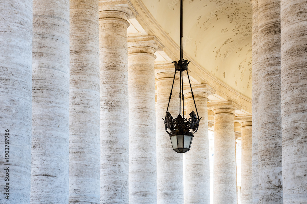 Naklejka premium Visitors walk through the colonnade at St Peters Square in Vatican. The tall columns create a grand space under the ceiling. A light fixture hangs down, adding to the design of the area.