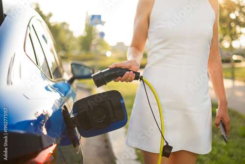 Woman connects a charging cable to her electric car in a bright, green area during the day.