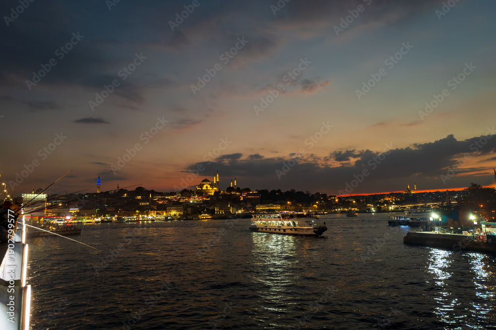 Fototapeta premium Anglers line the Galata Bridge at dusk as illuminated Suleymaniye Camii crowns the Eminonu skyline, with ferries crossing the Golden Horn under a warm after-sunset sky