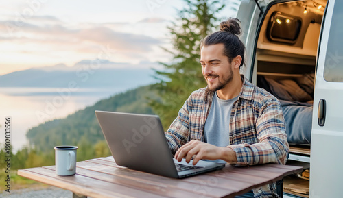 A man freelancer sits at table and works on his laptop near a travel van in the backdrop of beautiful nature during his weekend in the wilderness. Remote work during a trip.