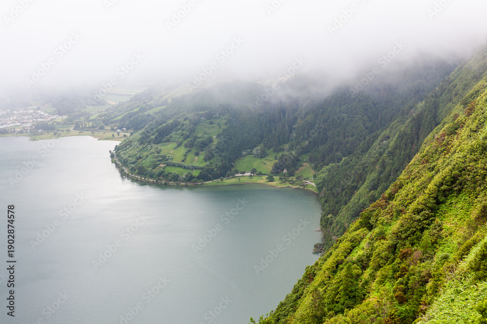 Fototapeta premium Lake surrounded by green hills and fog at Sete Cidades, San Miguel, Azores. Lush forested landscape in the background. A volcanic crater lake in Portugal.