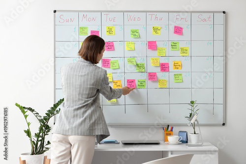 Young businesswoman sticking notes with plans on whiteboard in office, back view