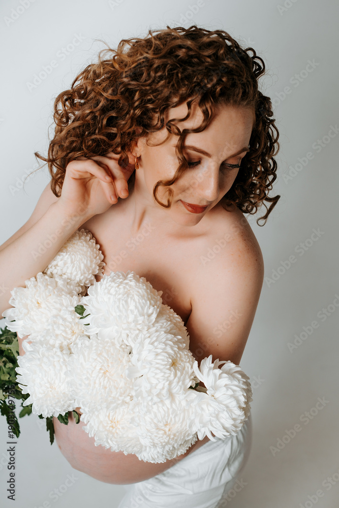 Fototapeta premium a pregnant woman with flowers on a white isolated background patting her belly and enjoying her pregnancy, a young girl with a big belly at home waiting for a baby, showing a heart with her hands
