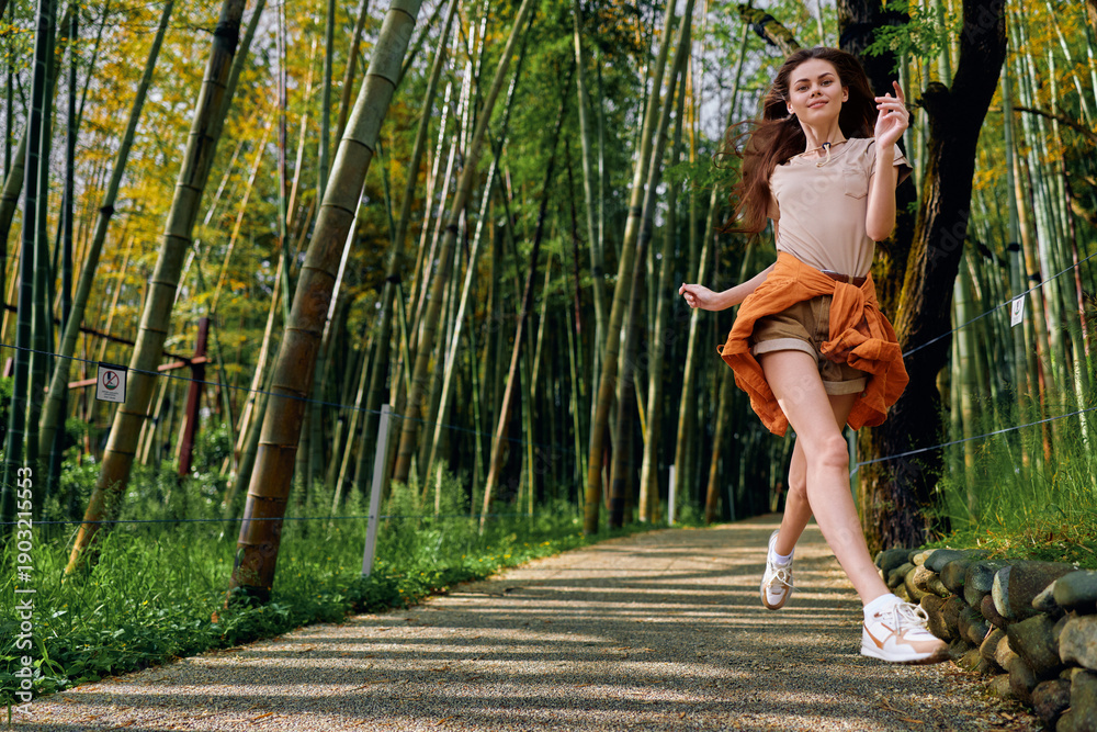Naklejka premium Woman jump along a bamboo path in a forest, casual summer outfit and smile, motion captured midair on a sunny outdoor walkway conveying joyful energy and carefree lifestyle.