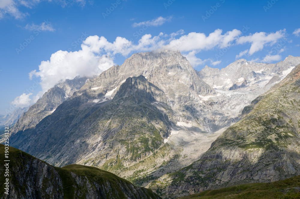 Obraz premium Imposing mountain peaks and rugged cliffs tower above a glacier-carved valley, with scattered clouds casting soft shadows on the rocky landscape under a bright blue sky.