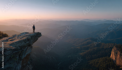Person standing on the edge of a rugged outdoor cliff overlooking a vast mountain landscape during sunset
