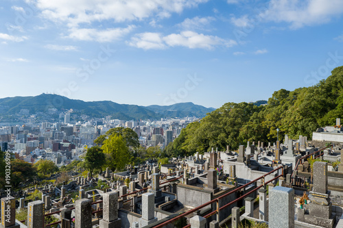 Photos Rows of stone graves on a terraced hillside cemetery overlook the urban skyline of Nagasaki, Japan, with distant green mountains and a clear blue sky