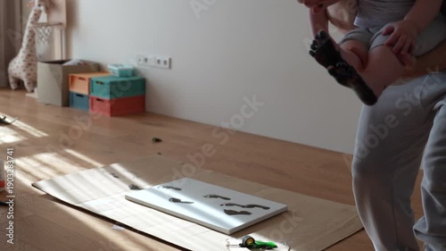 Selfie UGC video of parents holding a baby and showing a paper with footprint painting. Black baby footprints visible on the sheet during a diy activity at home.
