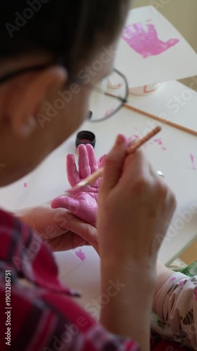 mother painting a toddler’s hand with a brush during a simple art activity at a table. close up view of adult hands and child’s hand. indoor home setting