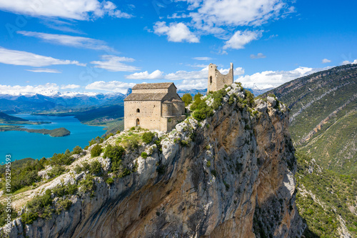 Historic stone castle and church rise above a steep rocky escarpment overlooking the brilliant turquoise waters of Mediano lake in Aragon, Spain. Crisp sunlight highlights the rugged limestone