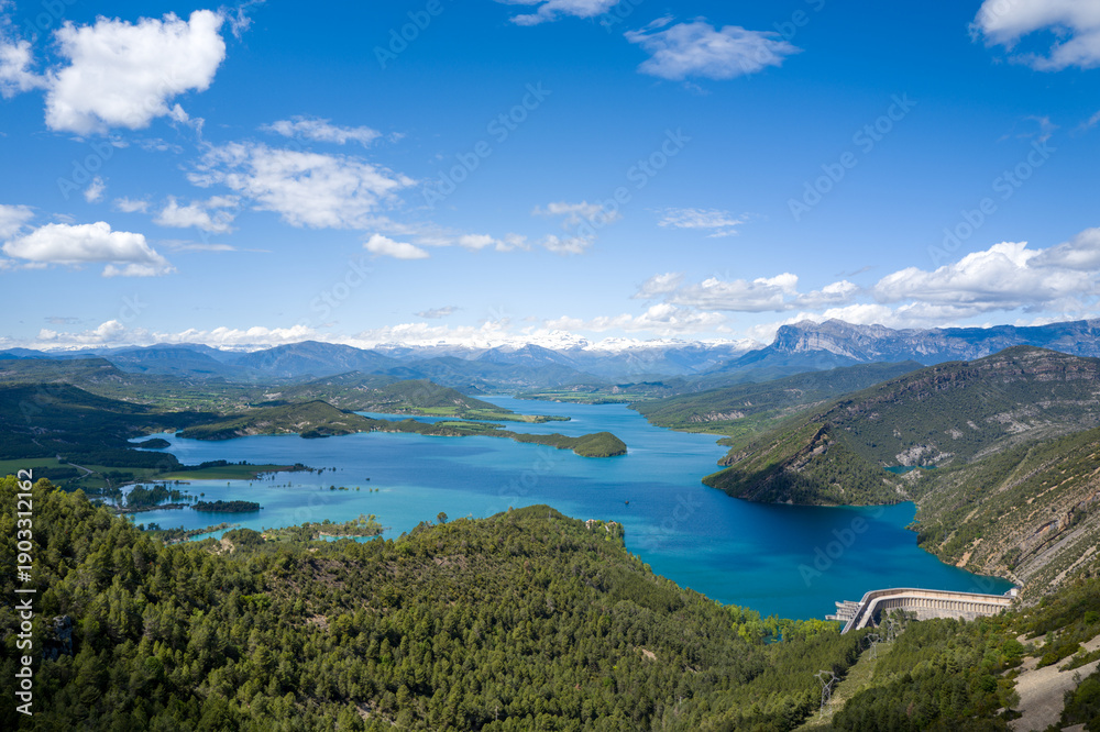 Fototapeta premium Expansive aerial landscape of Mediano Lake in Aragon, Spain, featuring turquoise water, lush green hills, and distant snow-capped Pyrenees mountains beneath a vivid blue sky with scattered clouds.