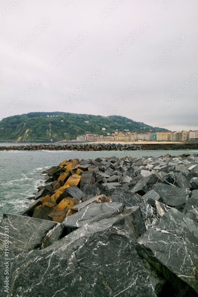 Fototapeta premium Vertical view of massive grey stones of a breakwater in the foreground, with the sea water, city skyline, and a prominent green hill under a cloudy sky in San Sebastian, Basque Country, Spain territor