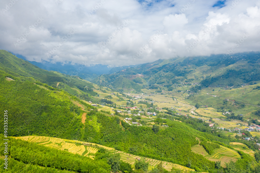 Fototapeta premium Wide panoramic scene of lush green hills and layered rice terraces surrounding a rural valley near Sapa, Vietnam. Cloud-filled skies cast soft light over the vibrant landscape and distant village.
