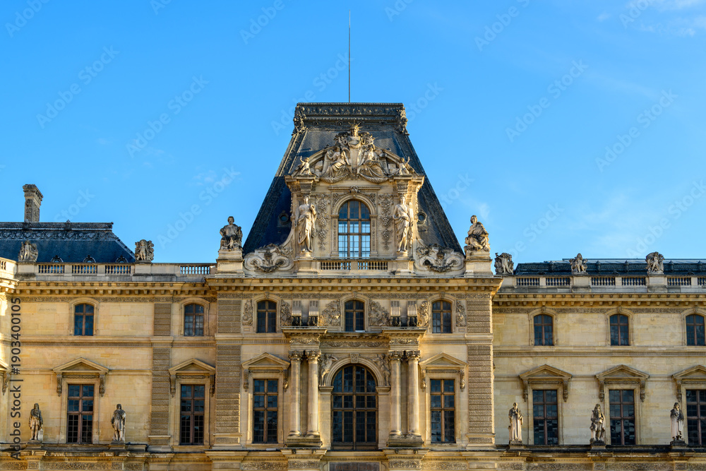 Obraz premium Symmetrical frontal view of the Louvre palace's central pavilion in Paris, France, featuring detailed classical statues and elaborate stone reliefs beneath a slate mansard roof. The historic facade is