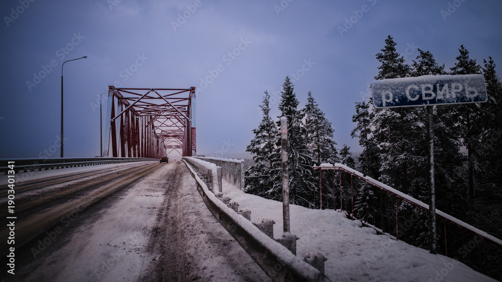 Fototapeta premium Snowy bridge crossing in northern Russia. Icy road leads through a red steel bridge surrounded by winter forest, with a road sign covered in snow and a cold, moody sky.