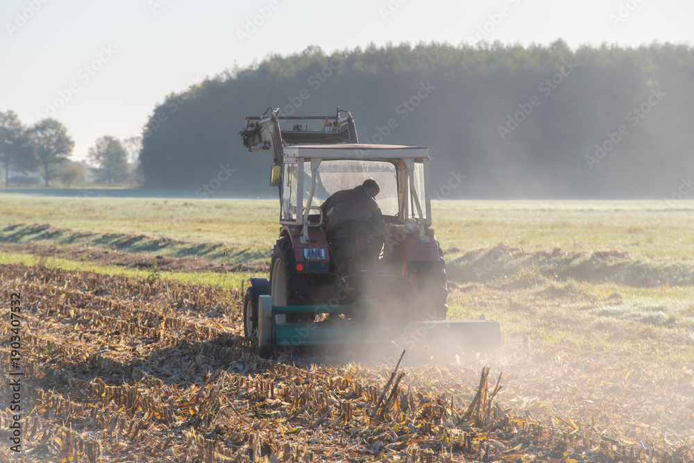 Fototapeta premium Traktor bei der Feldarbeit nach der Maisernte, Landwirtschaftliche Feldarbeit nach der Maisernte mit Traktor und Erntemaschine in ländlicher Landschaft.