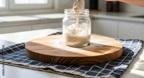 Hand pours floury mixture into glass jar, on wood board, kitchen, with sunlight
