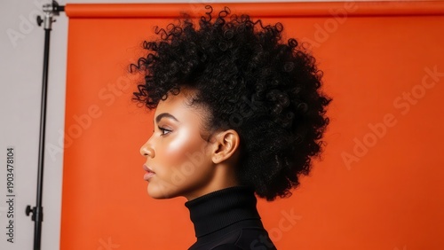 Profile view of a young woman with voluminous afro hair and glowing skin, wearing a black turtleneck against a vibrant orange studio background.
