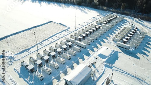Aerial view of a large battery energy storage facility in winter, showing containerized power units casting long shadows on snow with forest surroundings and smooth orbital motion.