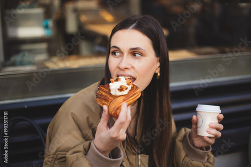 Young woman enjoying delicious pastry and coffee at outdoor cafe, happy girl eating sweet croissant or bun and holding paper cup, urban lifestyle and breakfast concept, street style portrait