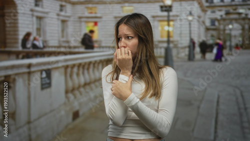 Photos Woman bites nails with hand to chest on crowded city street beside stone building and lamppost; anxiety vulnerability