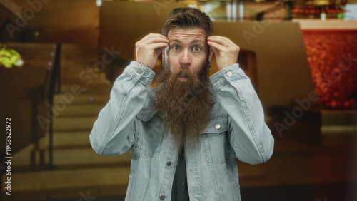 Φωτογραφία Bearded man pulls forehead skin with both hands in a modern hotel lobby staircase setting; stress tension