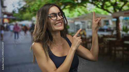 Woman wearing round glasses smirking and pointing index finger on busy street terrace at restaurant seating; playfulness.