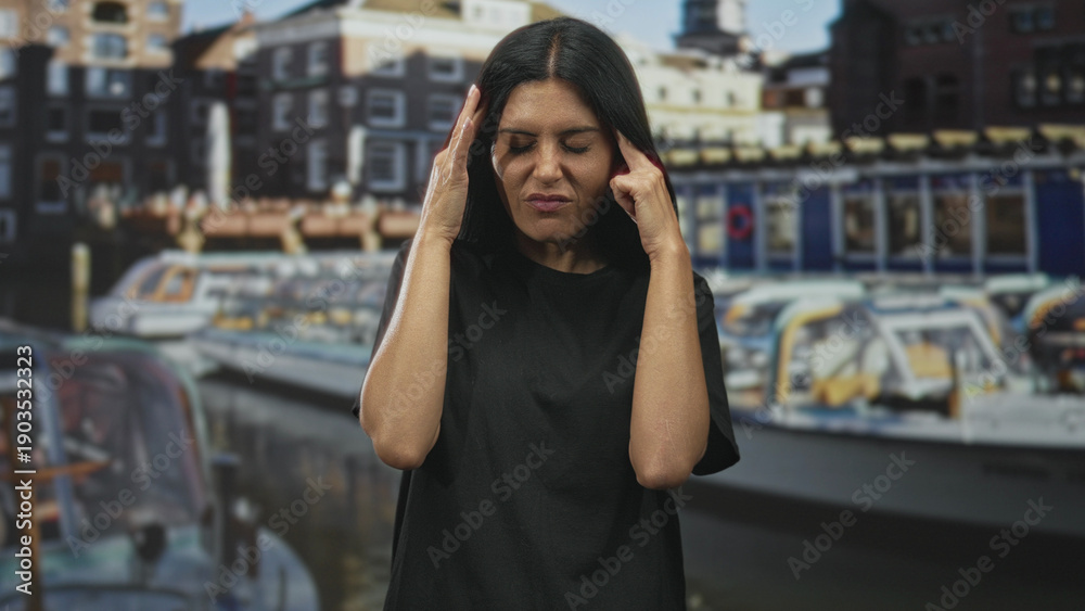 Fototapeta premium Woman pressing temples with hands, eyes closed and grimacing, visible forehead and fingers, wearing a black t shirt by canal boats on a street with buildings; stress fatigue tension.
