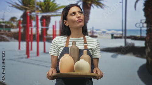 Canvas Print Woman artisan holding tray of ceramic vases with hands, looking sideways at a beach promenade; craftsmanship calm