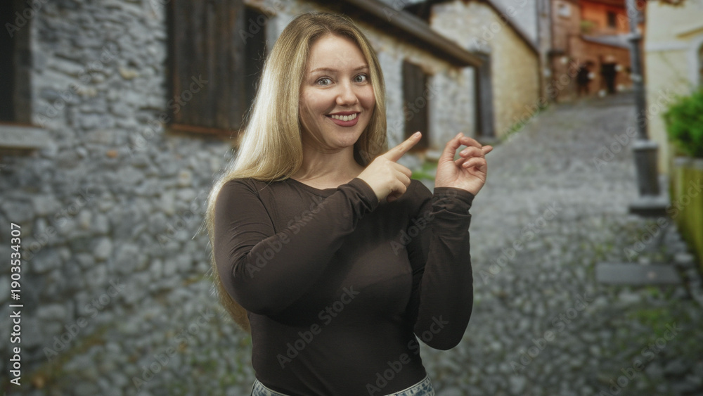 Fototapeta premium Woman smiling points both index fingers to the right while wearing a black shirt on a cobblestone street with stone walls; joy.