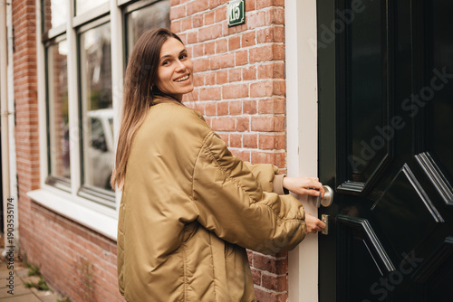 Quadro em tela Happy brunette woman in gold bomber jacket locking or open red brick house door,