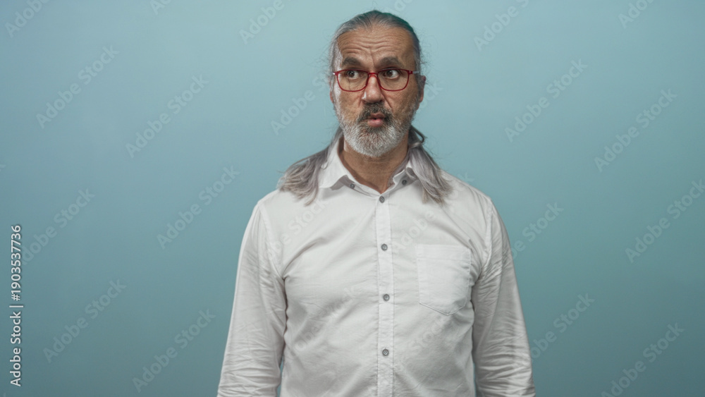 Obraz premium Man with long gray hair and beard wearing red glasses and white shirt purses lips in light blue studio; surprise and quiet introspection.