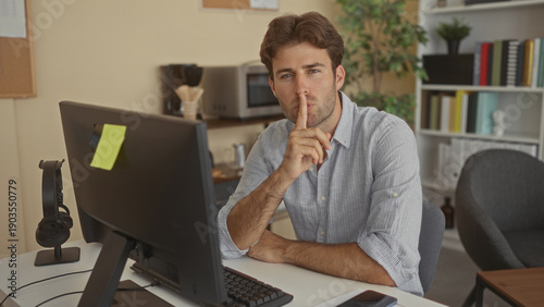Fotografie Man at desktop computer, finger to lips for silence in office building with headphones on stand, sticky note on monitor and bookshelf in view; silence focus