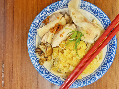 Top View of Chicken Noodle Bowl with Wontons, Mushrooms, and Red Chopsticks on Wooden Table