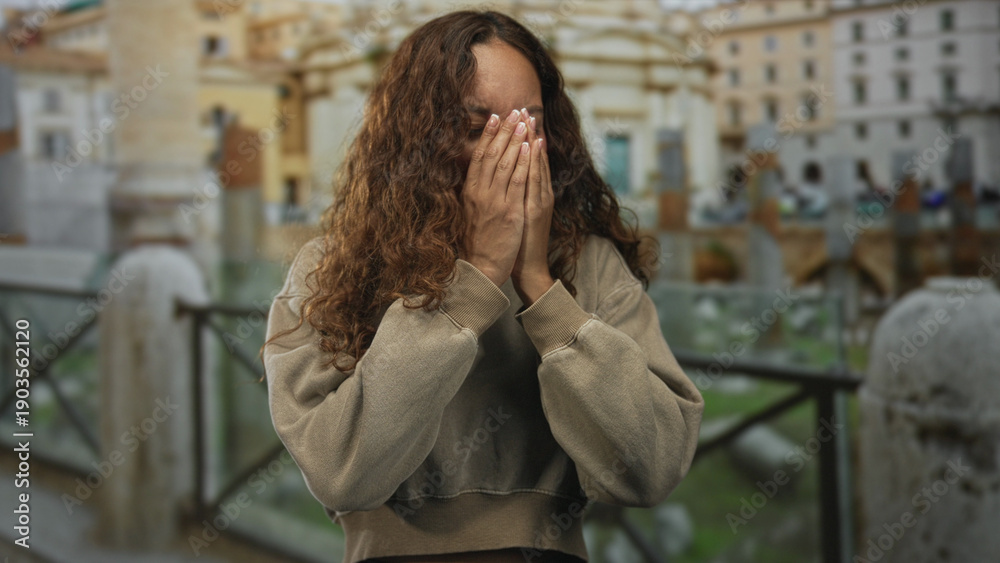 Fototapeta premium Young woman covering face with hands at ancient ruins building, eyes closed and bowed posture; sadness reflection solitude.