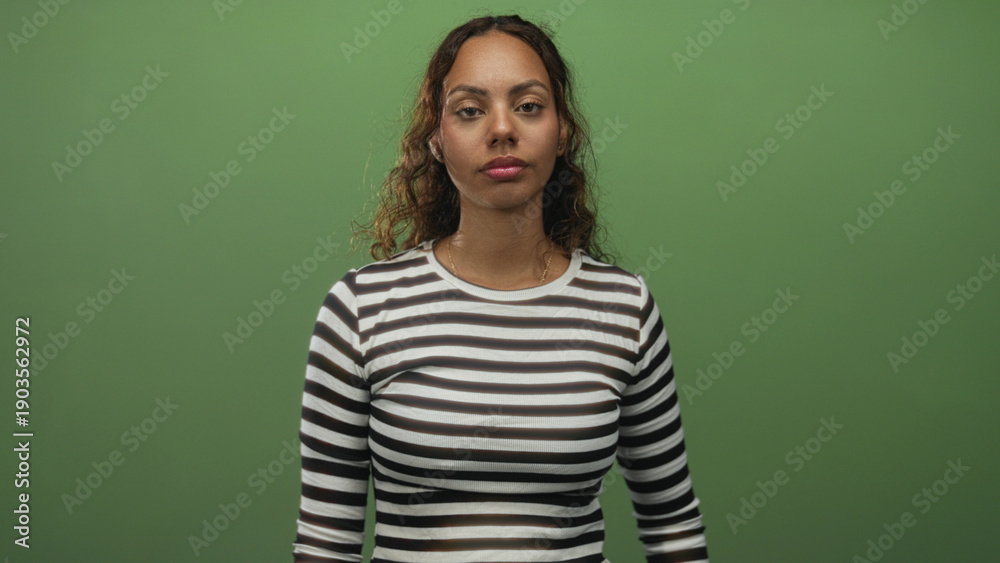 Fototapeta premium Woman points index finger upward in studio wearing striped shirt against green backdrop; confidence focus.