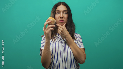 Fotografie Young hispanic brunette woman holding a muffin while touching chin with hand in teal studio, striped shirt visible; doubt