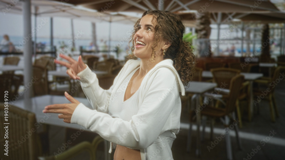 Fototapeta premium Young woman with curly hair smiles and gestures with hands, showing navel piercing in an outdoor seaside cafe by the boardwalk; joyful carefree.